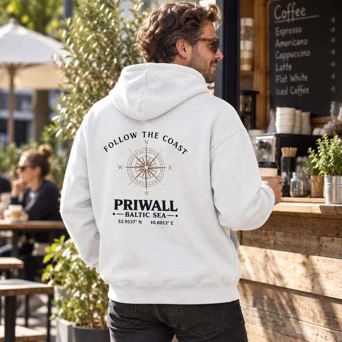 Mann im Bio Oversize Hoodie mit hellem Follow the Coast Rückenmotiv an einem Kaffeestand