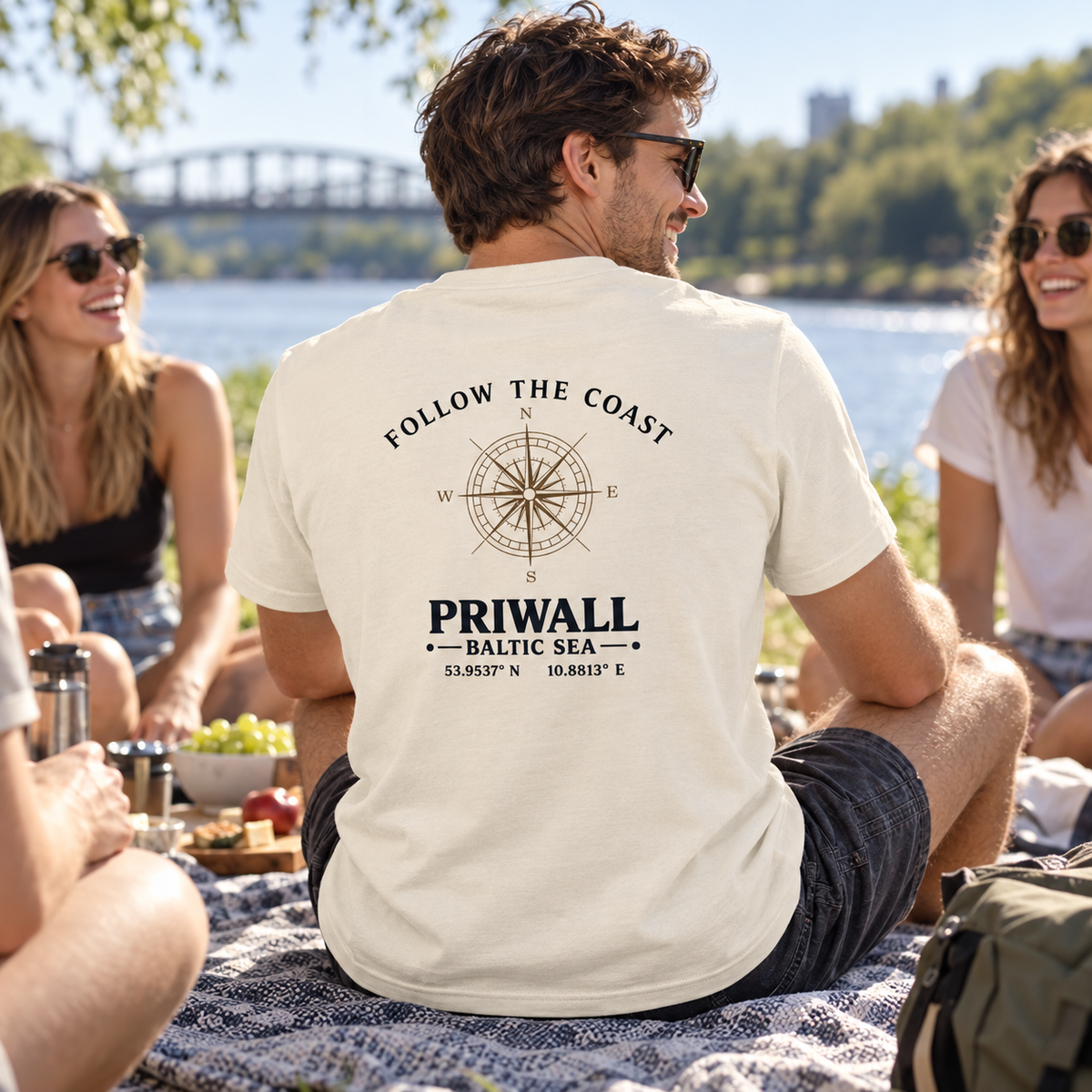 Mann im Bio T-Shirt mit hellem Follow the Coast Rückenmotiv bei einem Picknick am Fluss