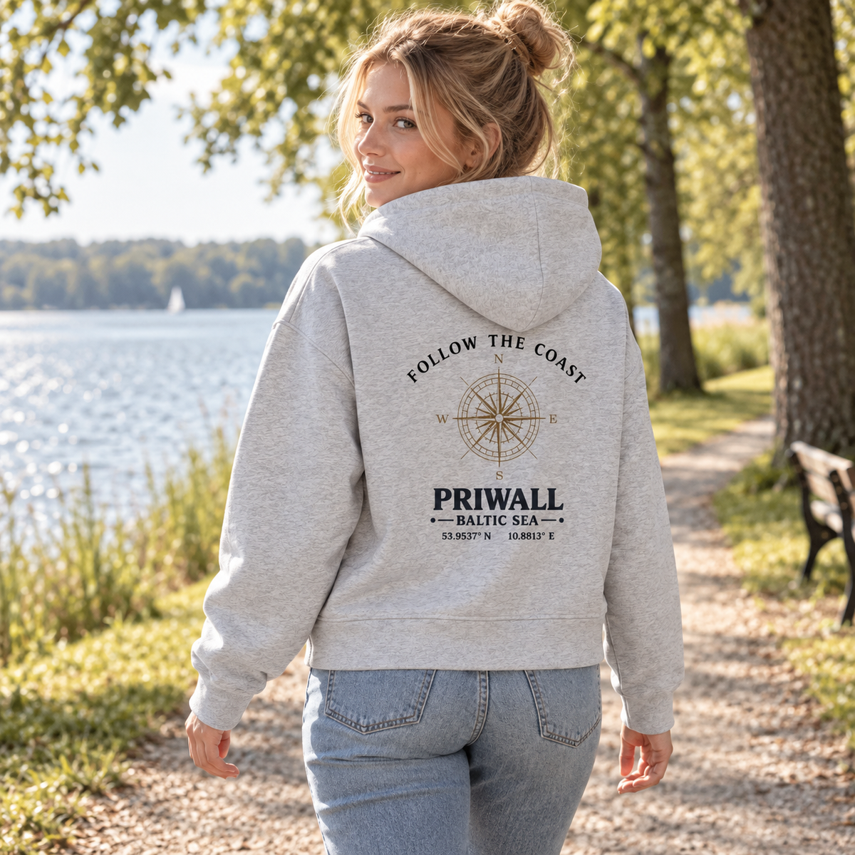 Frau im Bio Boxcut Hoodie mit hellem Follow the Coast Rückenmotiv bei einem Spaziergang am See