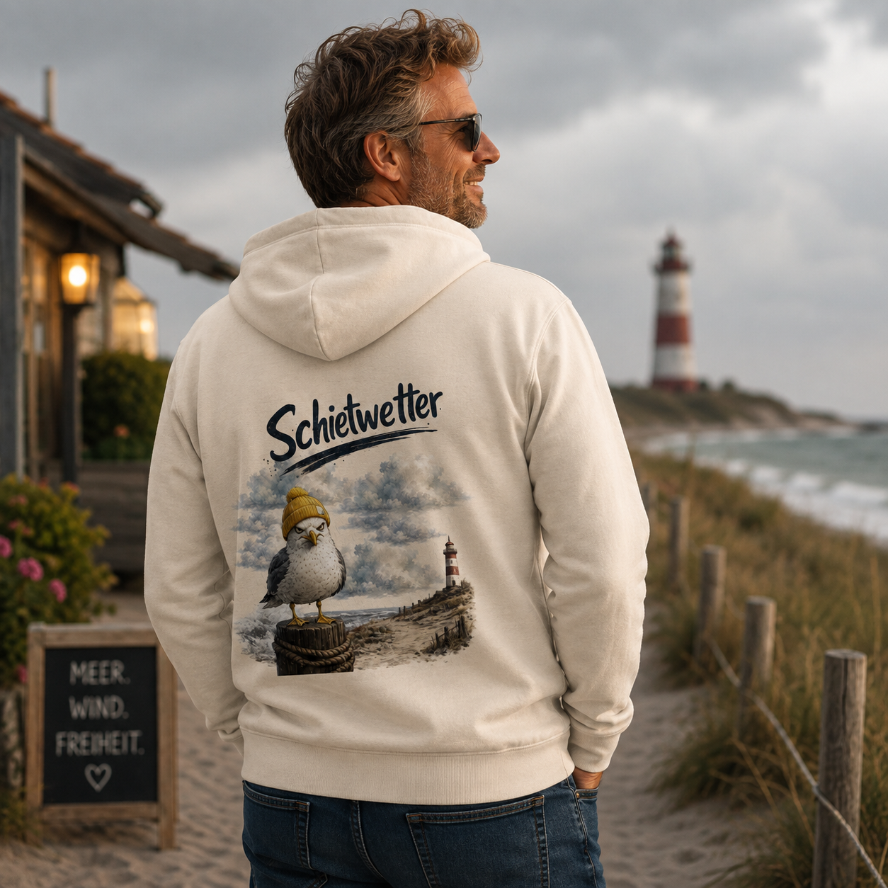 Mann trägt die Nomavé Bio Kapuzenjacke mit großem „Schietwetter“-Rückenprint und frecher Möwe mit gelber Mütze auf einem Dünenweg an der Ostsee bei bewölktem Himmel.