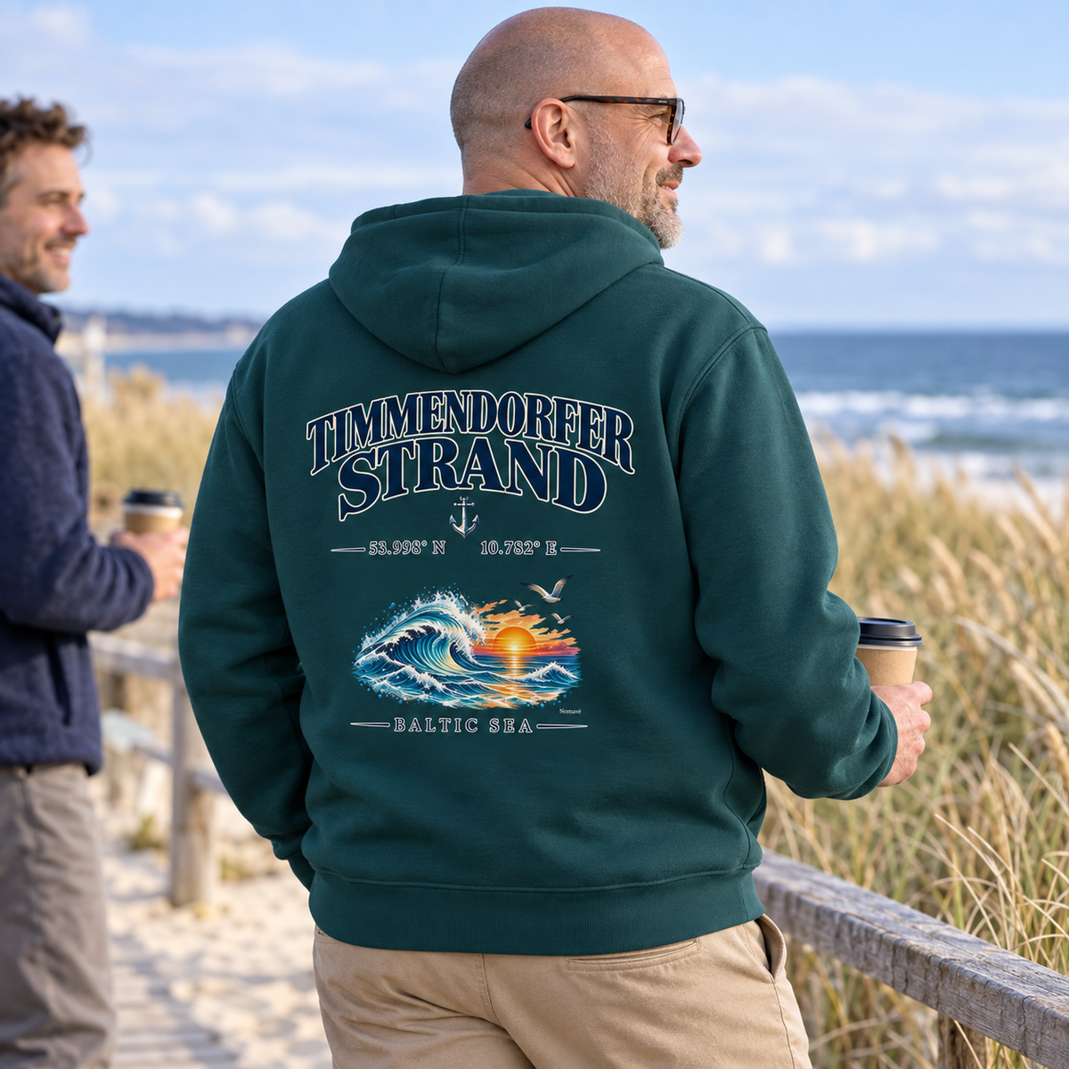 Mann im Bio-Hoodie in Glazed Green mit Timmendorfer Strand – Baltic Sea Rückenprint beim Kaffee am Strandweg.