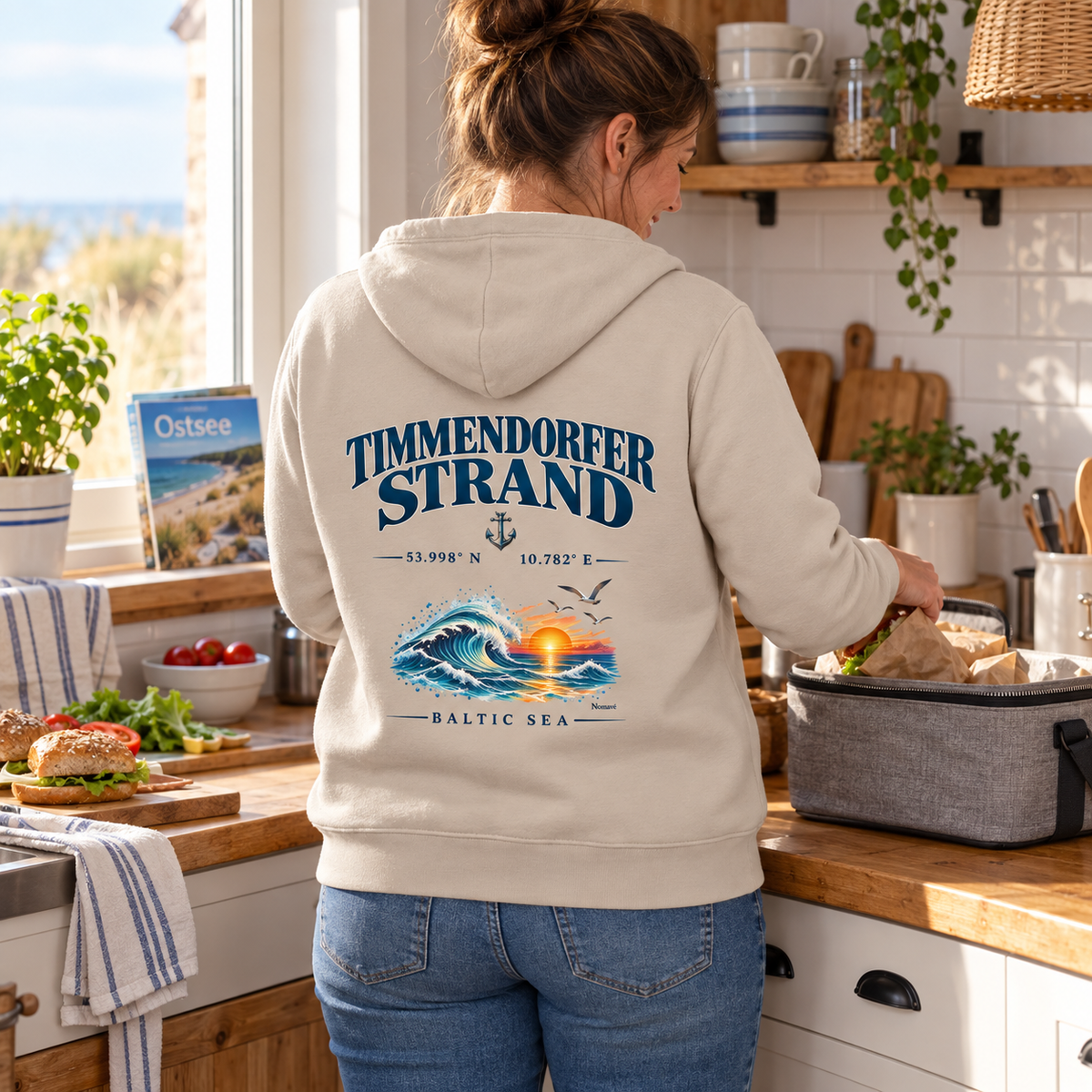 Frau im Bio-Hoodie in Desert Dust mit Baltic Sea Backprint beim Packen eines Picknicks in einer Ferienhausküche.