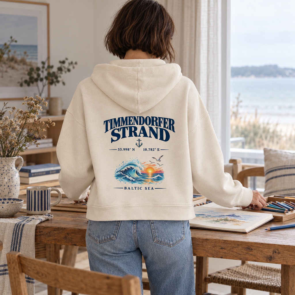 Frau im Damen Bio-Boxcut-Hoodie in Natural Raw mit Timmendorfer Strand – Baltic Sea Rückenprint beim Zeichnen in einer Ferienwohnung.