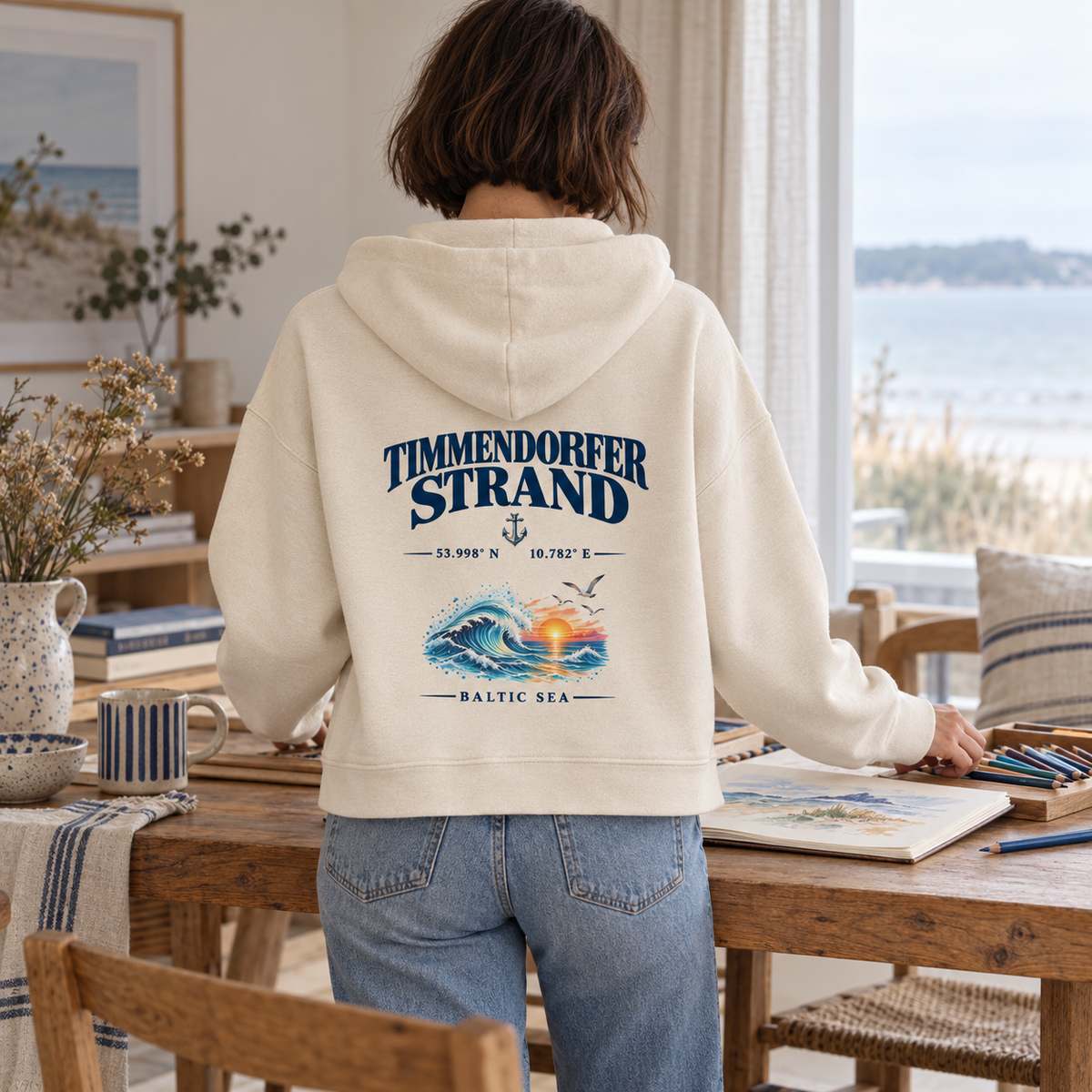 Frau im Damen Bio-Boxcut-Hoodie in Natural Raw mit Timmendorfer Strand – Baltic Sea Rückenprint beim Zeichnen in einer Ferienwohnung.