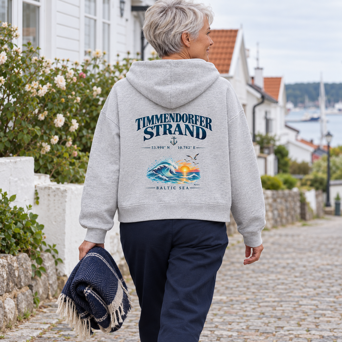 Frau im Damen Bio-Boxcut-Hoodie in Cool Heather Grey mit Timmendorfer Strand – Baltic Sea Rückenprint beim Spaziergang mit einer Picknickdecke durch einen Küstenort.
