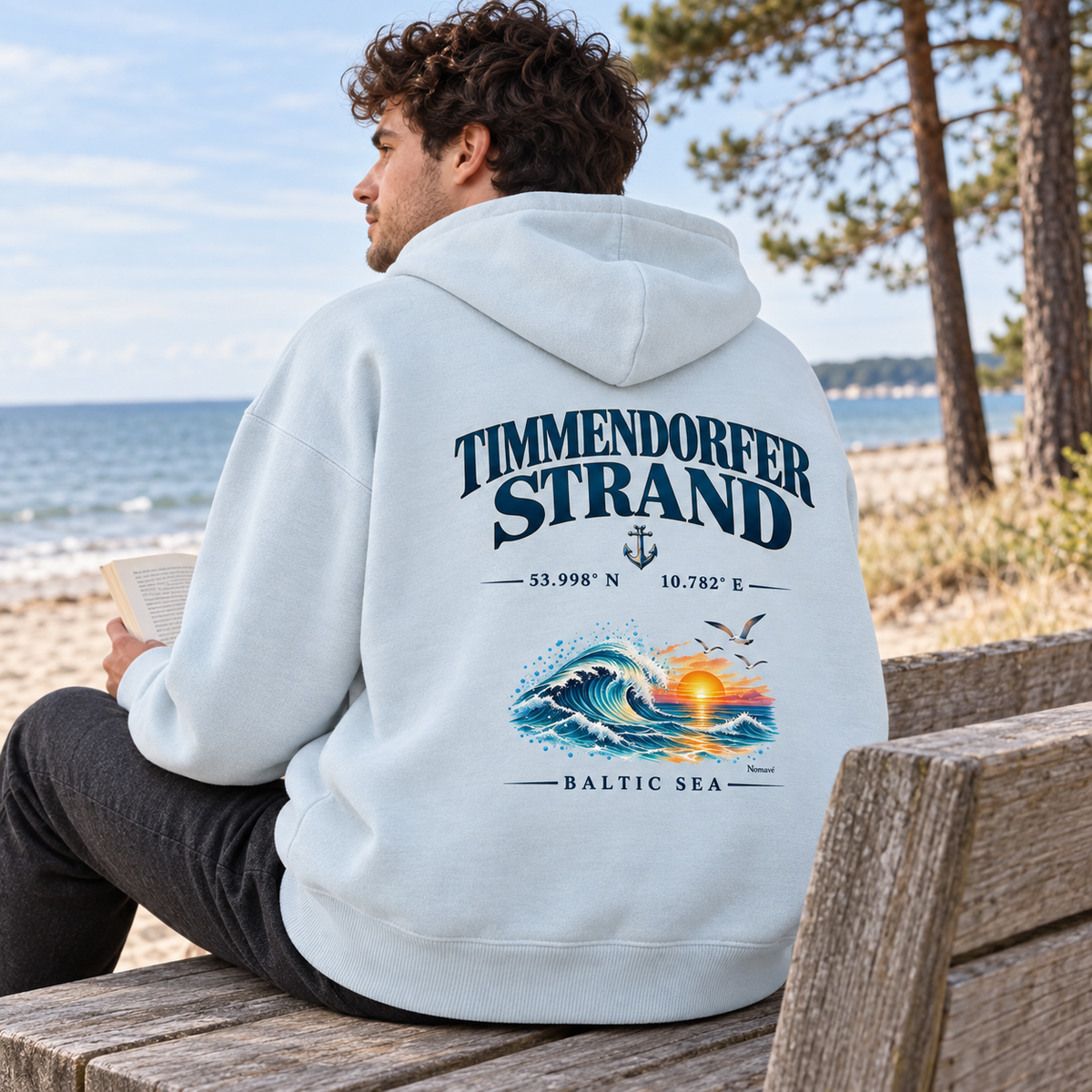 Mann im Bio-Oversize-Hoodie in Blue Ice mit Timmendorfer Strand – Baltic Sea Rückenprint beim Lesen auf einer Strandbank.