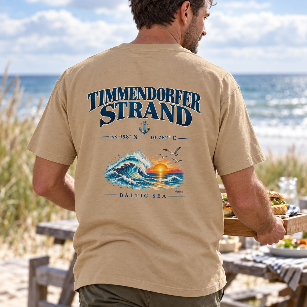 Mann im Bio-Heavy-T-Shirt in Latte mit Timmendorfer Strand – Baltic Sea Rückenprint bei einem Picknick am Meer.