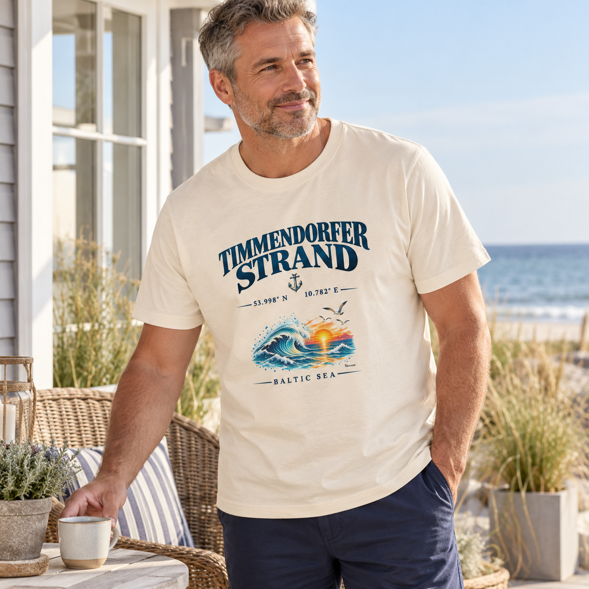 Mann im Unisex Bio-Heavy-T-Shirt in Natural Raw mit Timmendorfer Strand – Baltic Sea Frontprint beim Kaffee auf einer Terrasse.