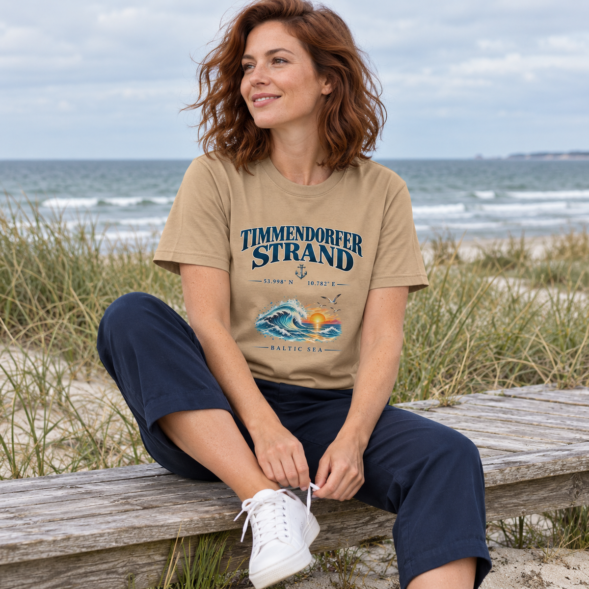 Frau im Unisex Bio-Heavy-T-Shirt in Latte mit Baltic Sea Frontprint beim Binden eines Sneakers auf einer Bank am Dünenweg.