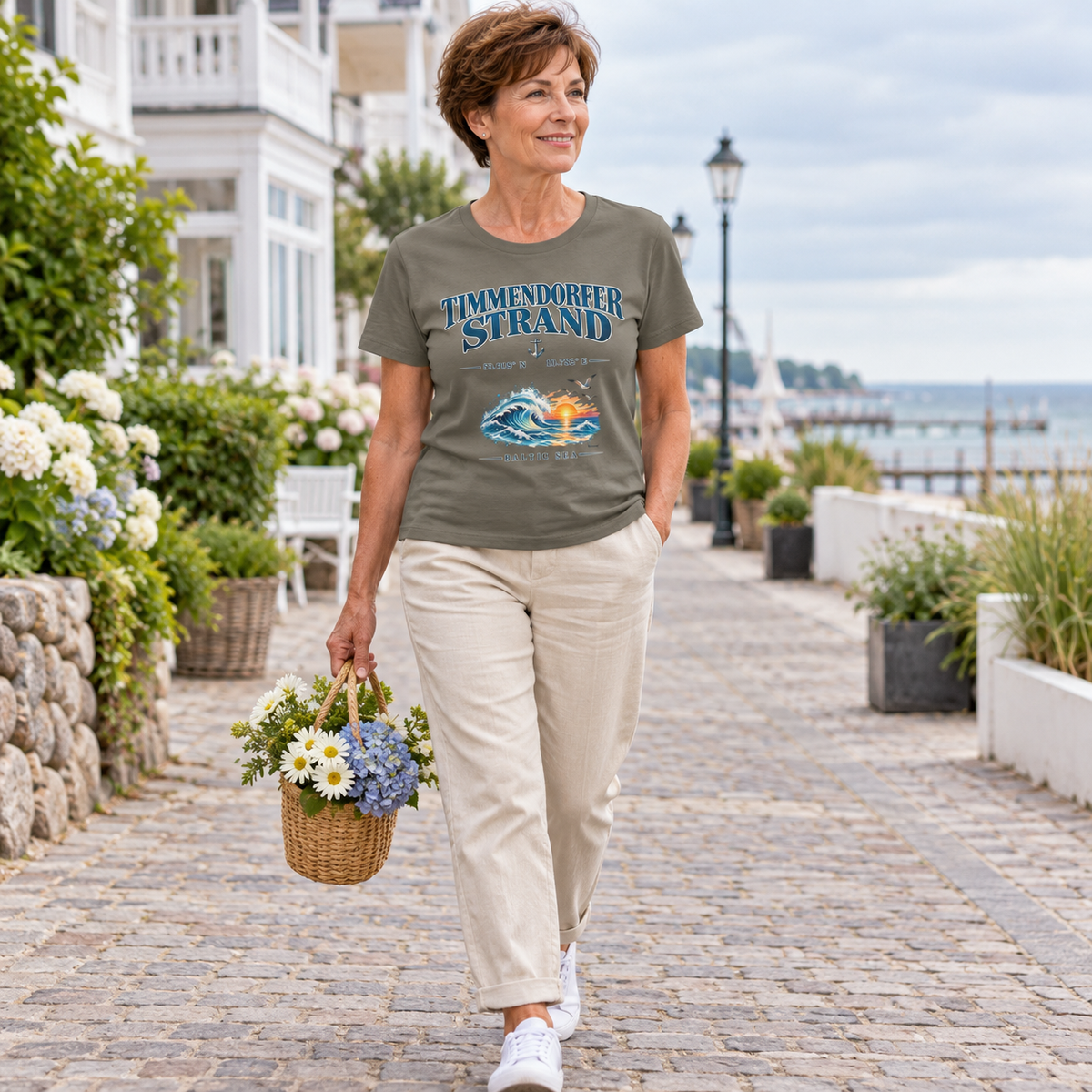Frau im Damen Bio-T-Shirt in Khaki mit Baltic Sea Frontprint beim Spaziergang mit einem Blumenkorb durch einen Küstenort.