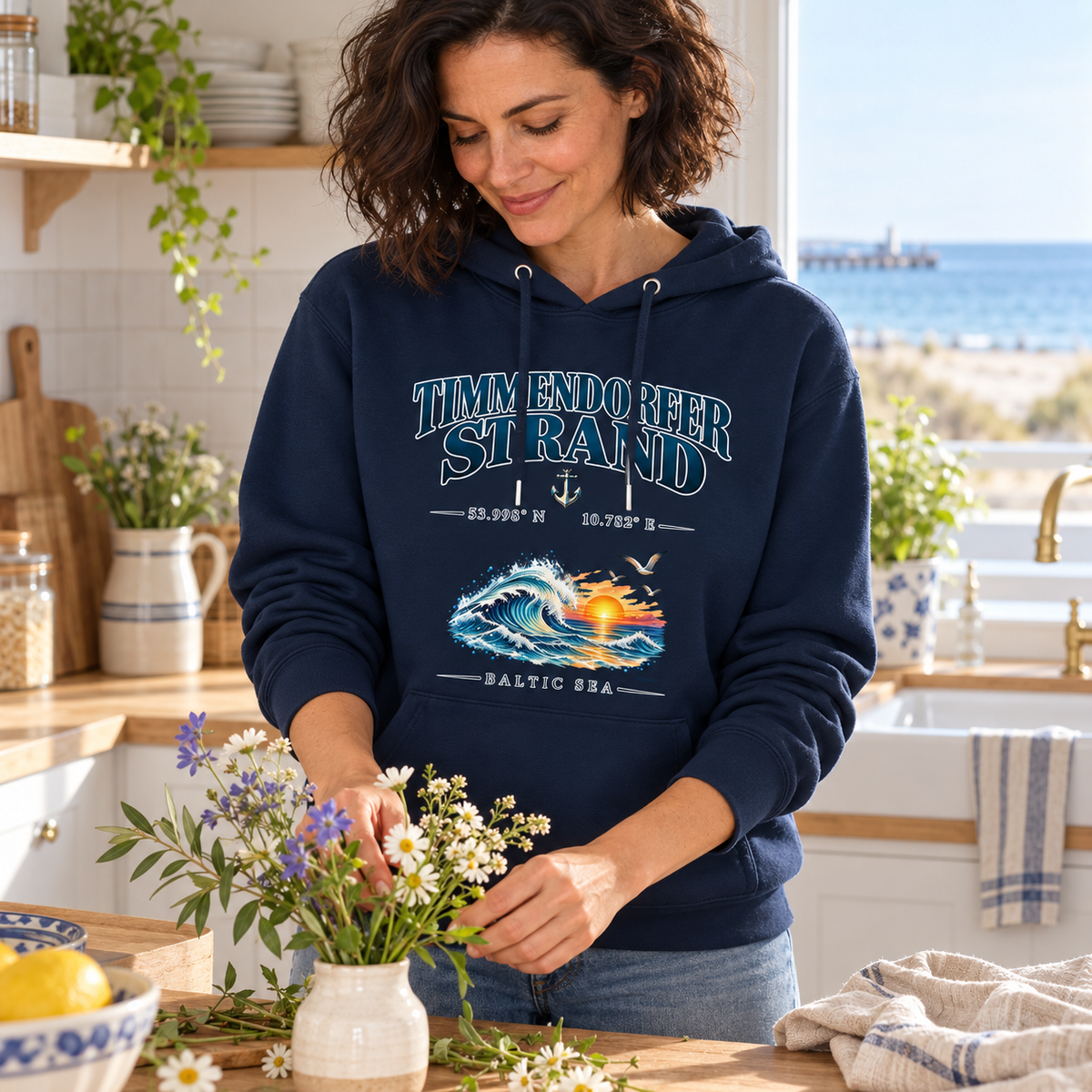 Unisex Bio-Hoodie in French Navy mit Timmendorfer-Strand–Baltic-Sea-Frontmotiv in heller Konturschrift: Frau arrangiert Blumen in einer Ferienhausküche.