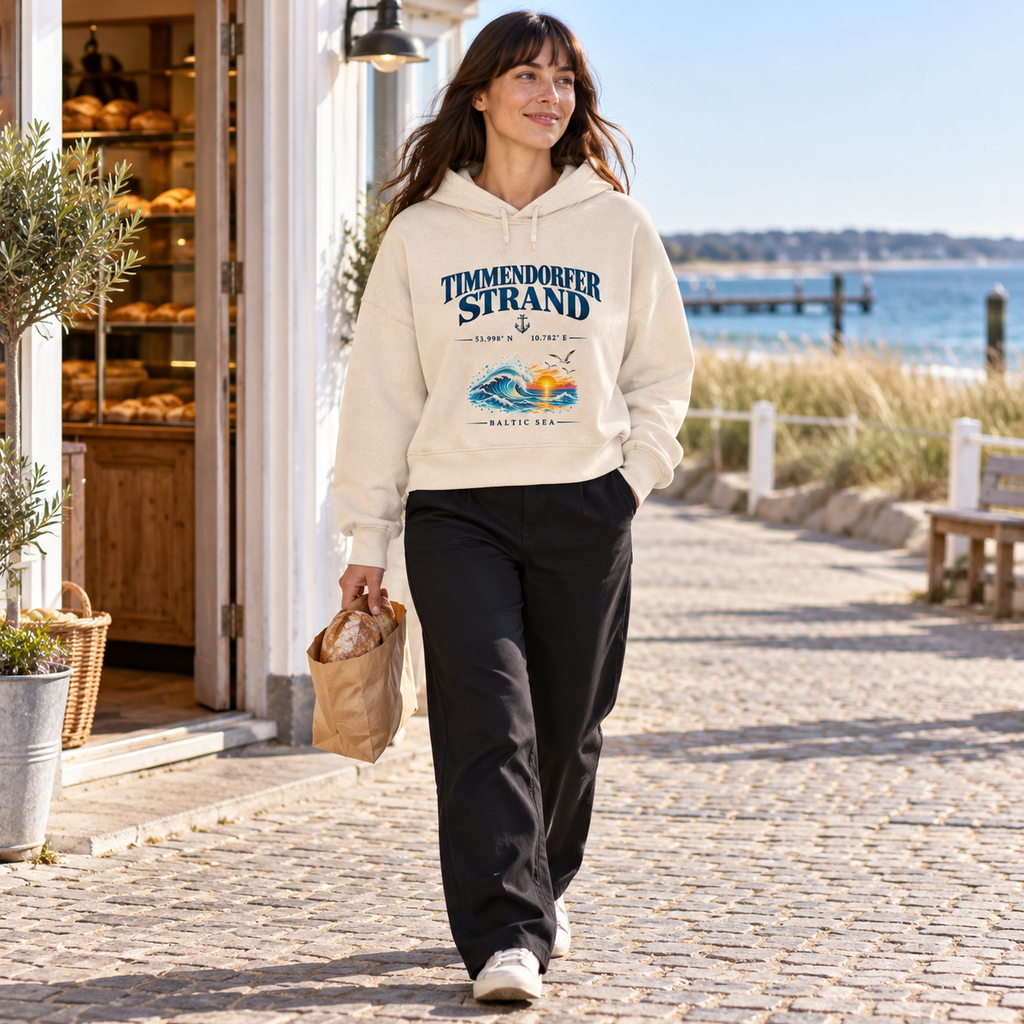 Damen Bio-Boxcut-Hoodie in Natural Raw mit Timmendorfer-Strand–Baltic-Sea-Frontmotiv: Frau geht mit einer Brottüte an einer Küstenbäckerei vorbei.