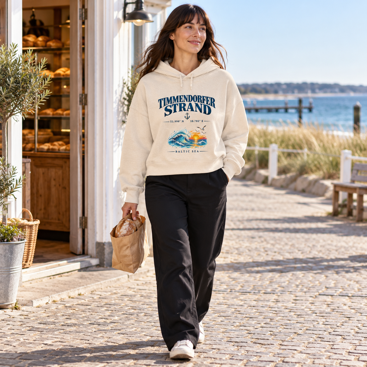 Damen Bio-Boxcut-Hoodie in Natural Raw mit Timmendorfer-Strand–Baltic-Sea-Frontmotiv: Frau geht mit einer Brottüte an einer Küstenbäckerei vorbei.