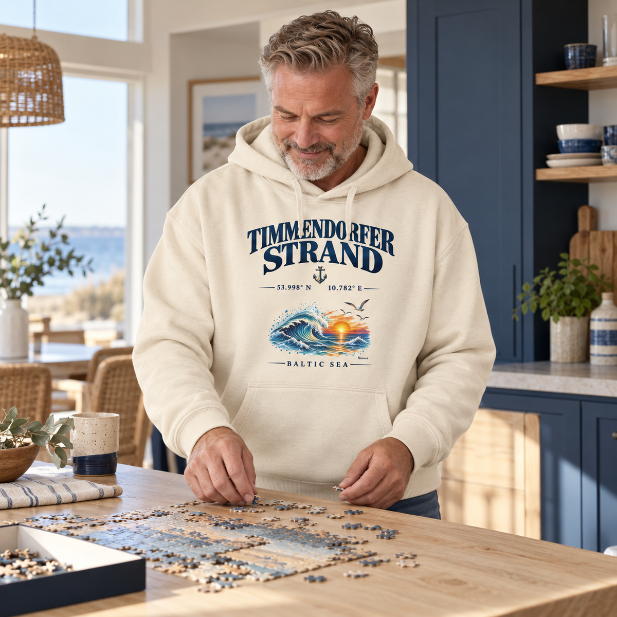 Unisex Bio-Oversize-Hoodie in Natural Raw mit Timmendorfer-Strand–Baltic-Sea-Frontmotiv: Mann legt ein Puzzle in einer Ferienhausküche.