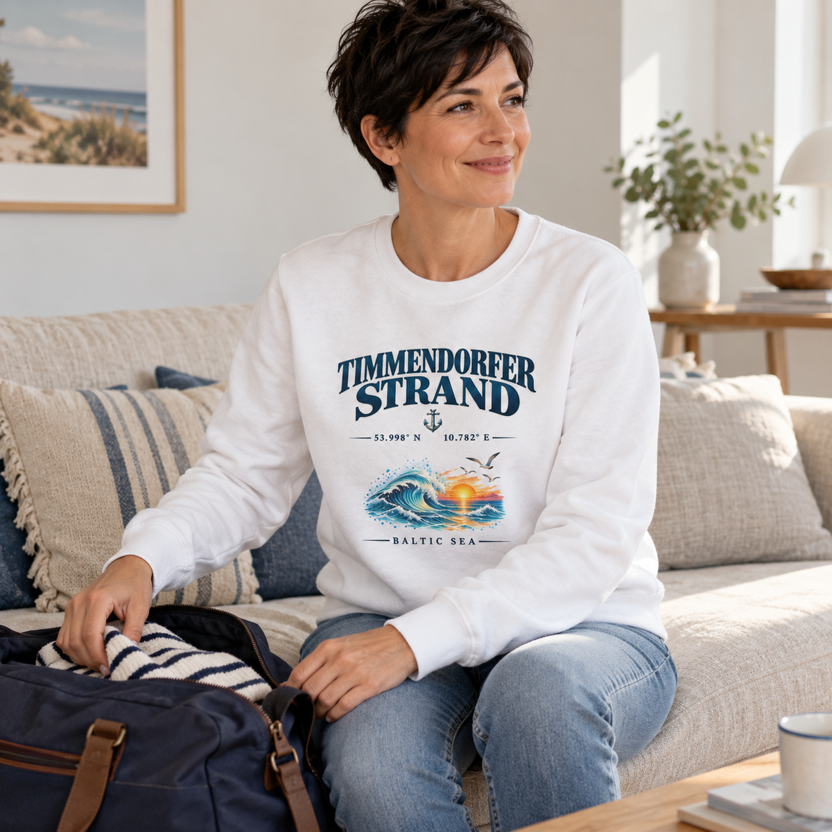 Unisex Bio-Sweatshirt in Weiß mit Timmendorfer-Strand–Baltic-Sea-Frontmotiv: Frau packt eine Reisetasche auf dem Sofa einer Ferienwohnung.