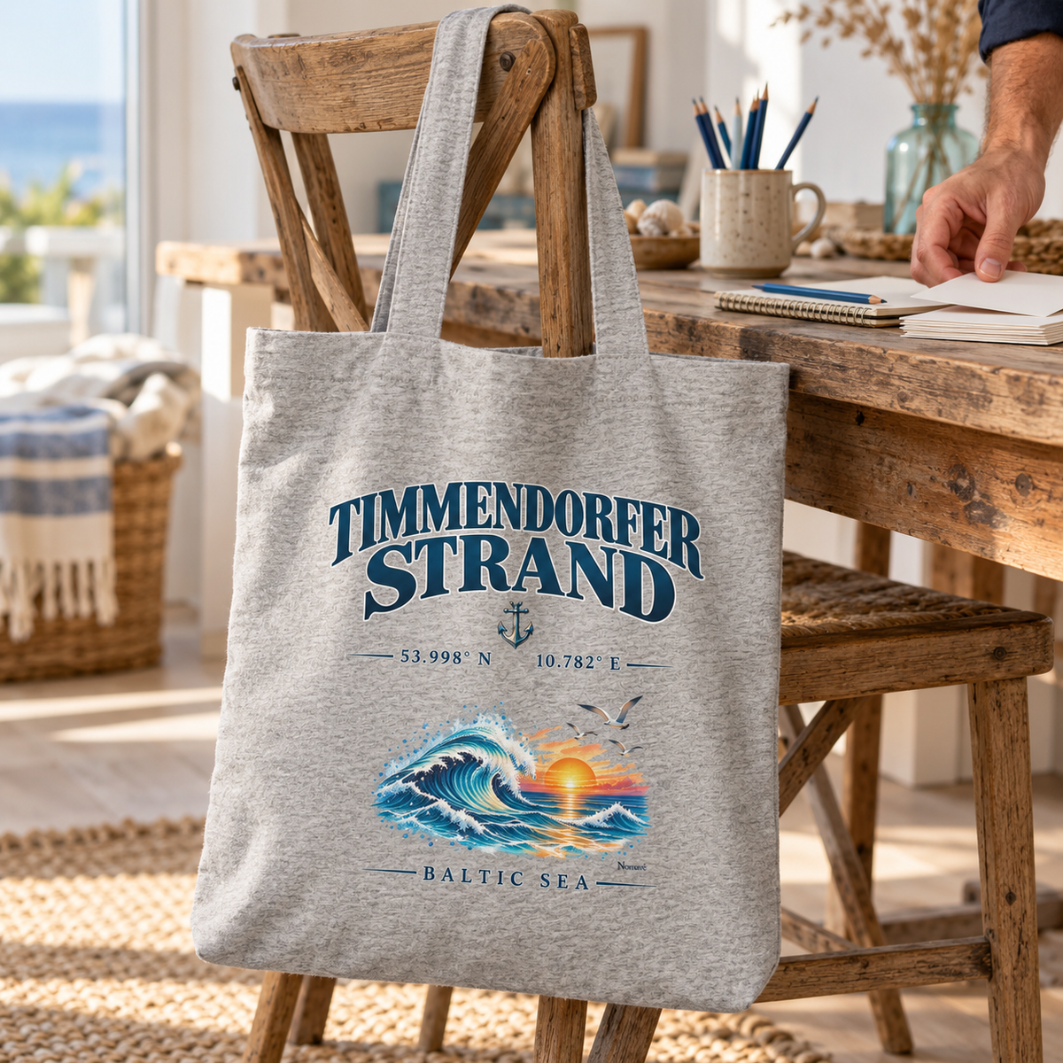 Einkaufsbeutel in Heather Grey mit Timmendorfer-Strand–Baltic-Sea-Frontmotiv: Tasche hängt an einem Holzstuhl neben einem kreativen Arbeitsplatz.
