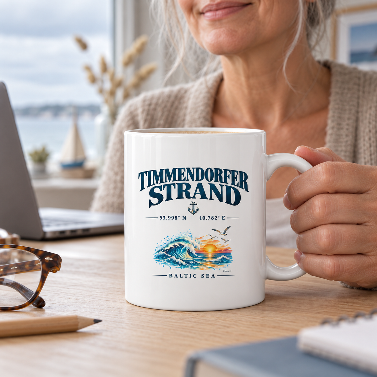 Glossy-Tasse 330 ml in Hochweiß mit Timmendorfer-Strand–Baltic-Sea-Motiv auf der Außenseite: Frau hält den C-Henkel am Homeoffice-Schreibtisch.