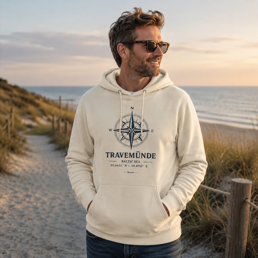 Ein Mann trägt einen cremefarbenen Nomavé Bio-Hoodie für Erwachsene mit dem Frontprint „Travemünde Baltic Sea“ und einer stilisierten Windrose. Er steht entspannt auf einem Dünenweg an der Ostsee im warmen Morgenlicht und blickt lächelnd auf das Meer.