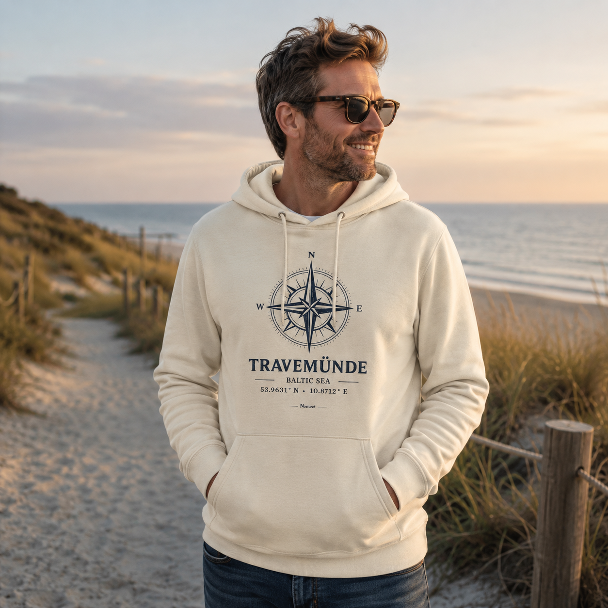 Ein Mann trägt einen cremefarbenen Nomavé Bio-Hoodie für Erwachsene mit dem Frontprint „Travemünde Baltic Sea“ und einer stilisierten Windrose. Er steht entspannt auf einem Dünenweg an der Ostsee im warmen Morgenlicht und blickt lächelnd auf das Meer.