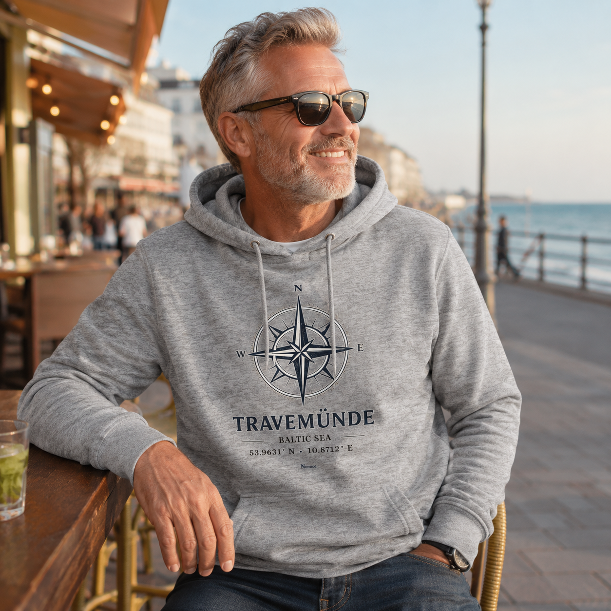 Ein Mann trägt einen grauen Nomavé Bio-Hoodie für Erwachsene mit dem Frontprint „Travemünde Baltic Sea“ und einer stilisierten Windrose. Er sitzt entspannt in einem Straßencafé an der Strandpromenade und blickt lächelnd aufs Meer – maritimes Lifestyle-Motiv mit Ostsee-Flair.