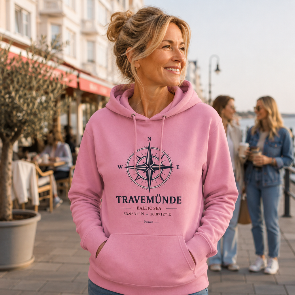 Eine Frau trägt einen rosa Nomavé Bio-Hoodie für Erwachsene mit dem Frontprint „Travemünde Baltic Sea“ und einer stilisierten Windrose. Sie steht lächelnd auf der Promenade an der Ostsee vor einem Café und genießt die maritime Atmosphäre an einem sonnigen Tag.