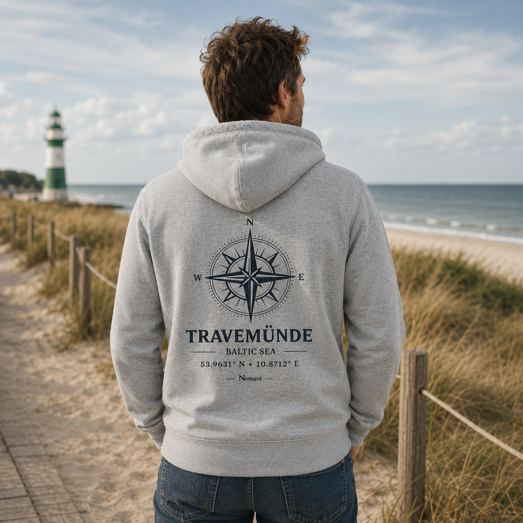 Junger Mann trägt eine hellgraue Nomavé Bio-Kapuzenjacke mit Reißverschluss am Ostseestrand. Die Rückenansicht zeigt den großen „Travemünde Baltic Sea“-Print mit Windrose und Koordinaten. Im Hintergrund sind Strand, Dünen, Meer und ein grün-weißer Leuchtturm zu sehen.