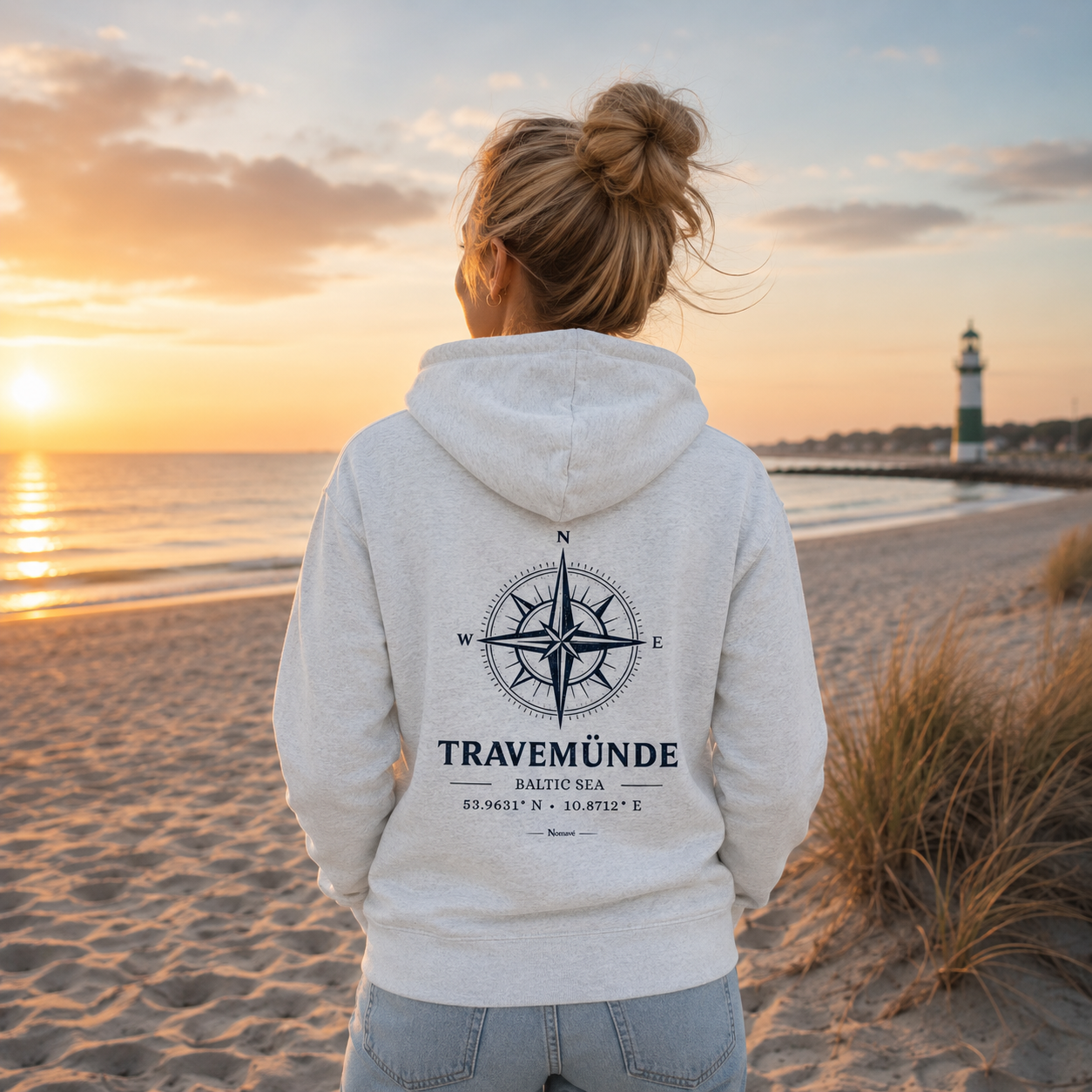 Rückenansicht einer Frau in einer Nomavé Bio-Kapuzenjacke mit Reißverschluss in Natural White. Der große „Travemünde Baltic Sea“-Kompassprint mit Koordinaten ist gut sichtbar. Im Hintergrund liegen Ostseestrand, Dünen, der grün-weiße Leuchtturm und warmes Morgenlicht.
