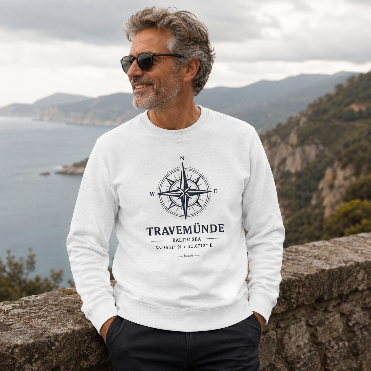 Ein Mann trägt ein weißes Nomavé Bio-Sweatshirt für Erwachsene mit dem Frontprint „Travemünde Baltic Sea“ und einer stilisierten Windrose. Er steht an einem Aussichtspunkt über der Küste und blickt lächelnd auf das Meer – maritimer Lifestyle-Look mit hochwertigem Outdoor-Ambiente.