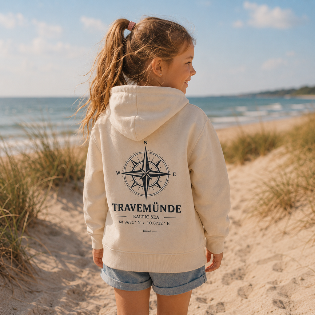 Mädchen trägt einen cremefarbenen Nomavé Kinder Bio-Hoodie mit großem „Travemünde Baltic Sea“-Kompassmotiv auf dem Rücken und steht zwischen den Dünen am Ostseestrand. Sommerliches Lifestylebild mit Meer und maritimer Urlaubsatmosphäre.
