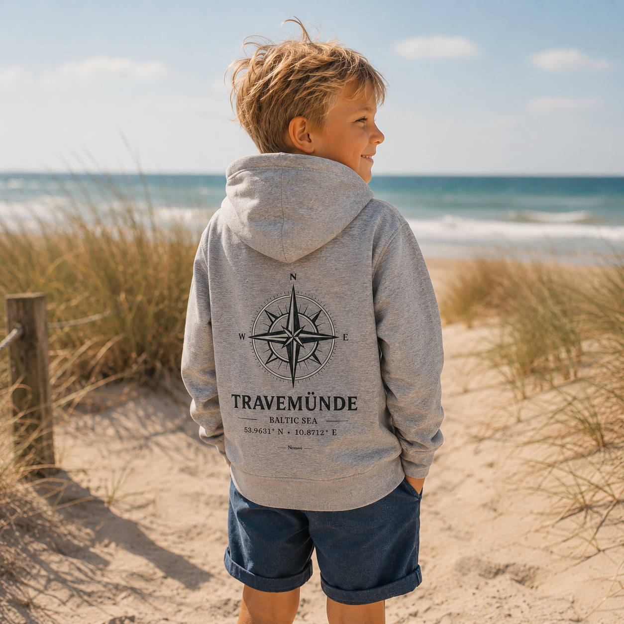 Kind trägt einen grauen Nomavé Kinder Bio-Hoodie mit großem „Travemünde Baltic Sea“-Kompassmotiv auf dem Rücken und blickt an einem sonnigen Tag über Dünen auf die Ostsee.