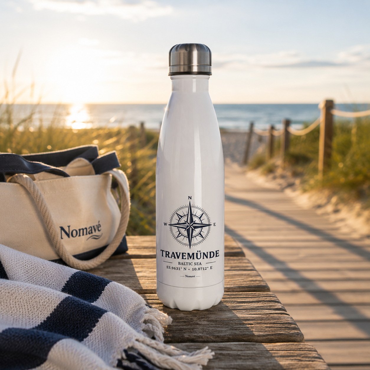 Weiße Edelstahl-Trinkflasche mit maritimem Kompassmotiv „Travemünde Baltic Sea“ von Nomavé auf einem Holztisch am Strandzugang zur Ostsee im sanften Morgenlicht. Umgeben von einer Strandtasche, einem Handtuch und den Dünen vermittelt die isolierte Trinkflasche das Gefühl von Sommer, Küstenurlaub und entspannten Tagen am Meer – ideal für Strand, Freizeit, Sport und Ausflüge.