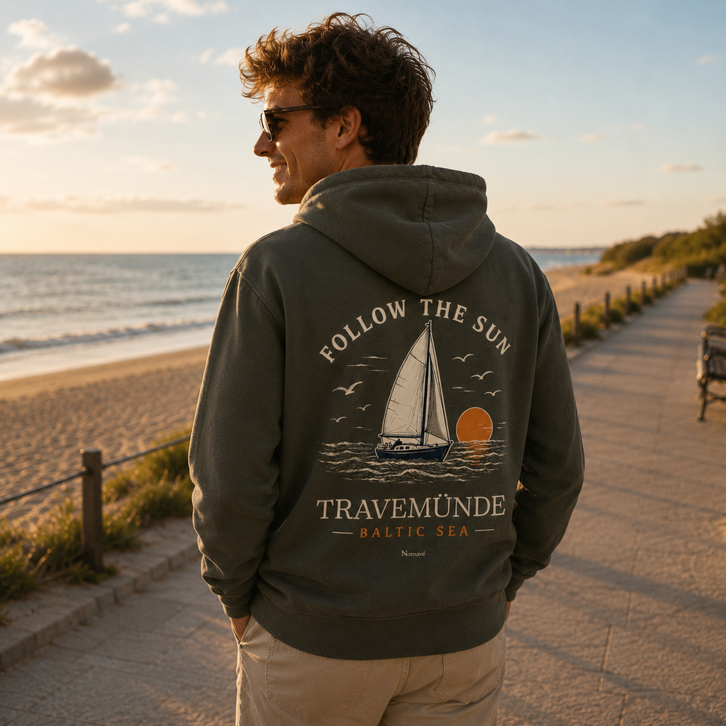 Mann trägt olivgrünen Travemünde Follow the Sun Hoodie mit großem Rückenprint und spaziert bei Sonnenuntergang an der Ostsee-Promenade